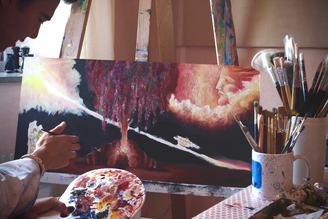 A wide painting on the easel in the studio: a column of light rises from a glowing point and spreads out in dark red, with a face in profile among the clouds on the right.