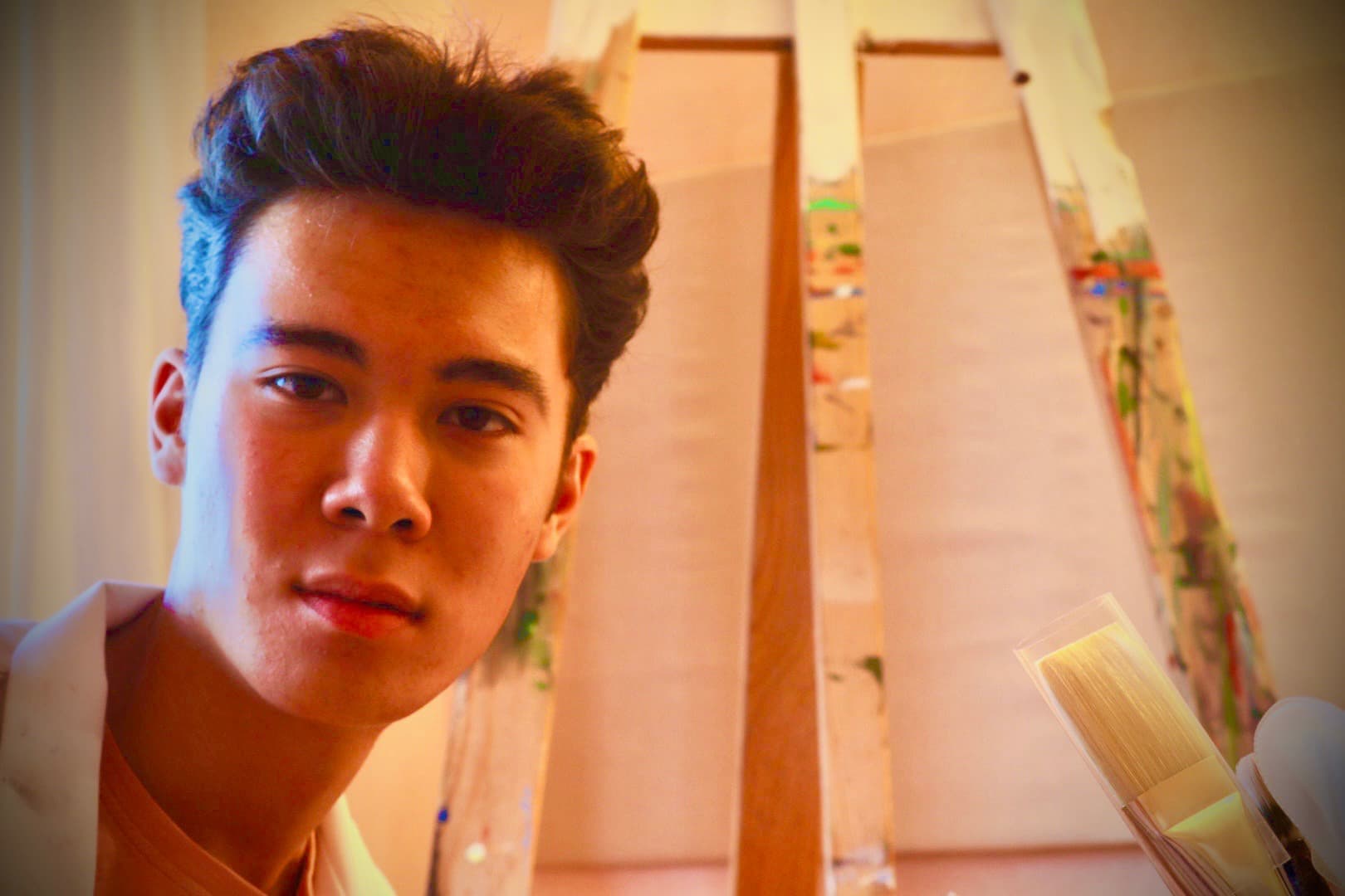 Isak in the studio, close up. Behind him is an easel with a pale canvas and paint-spattered wooden strips, and he is holding a wide brush.