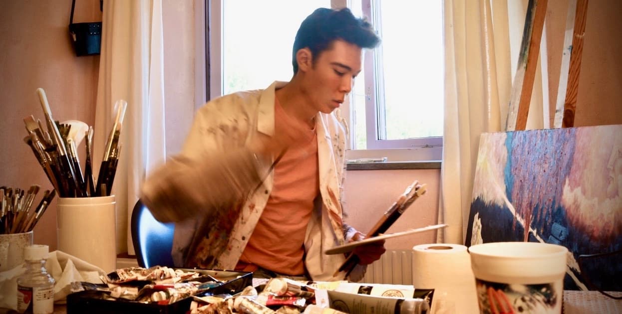 Isak working at a table covered with paint tubes, brushes and a palette. A window is behind him and a painting leans beside him.